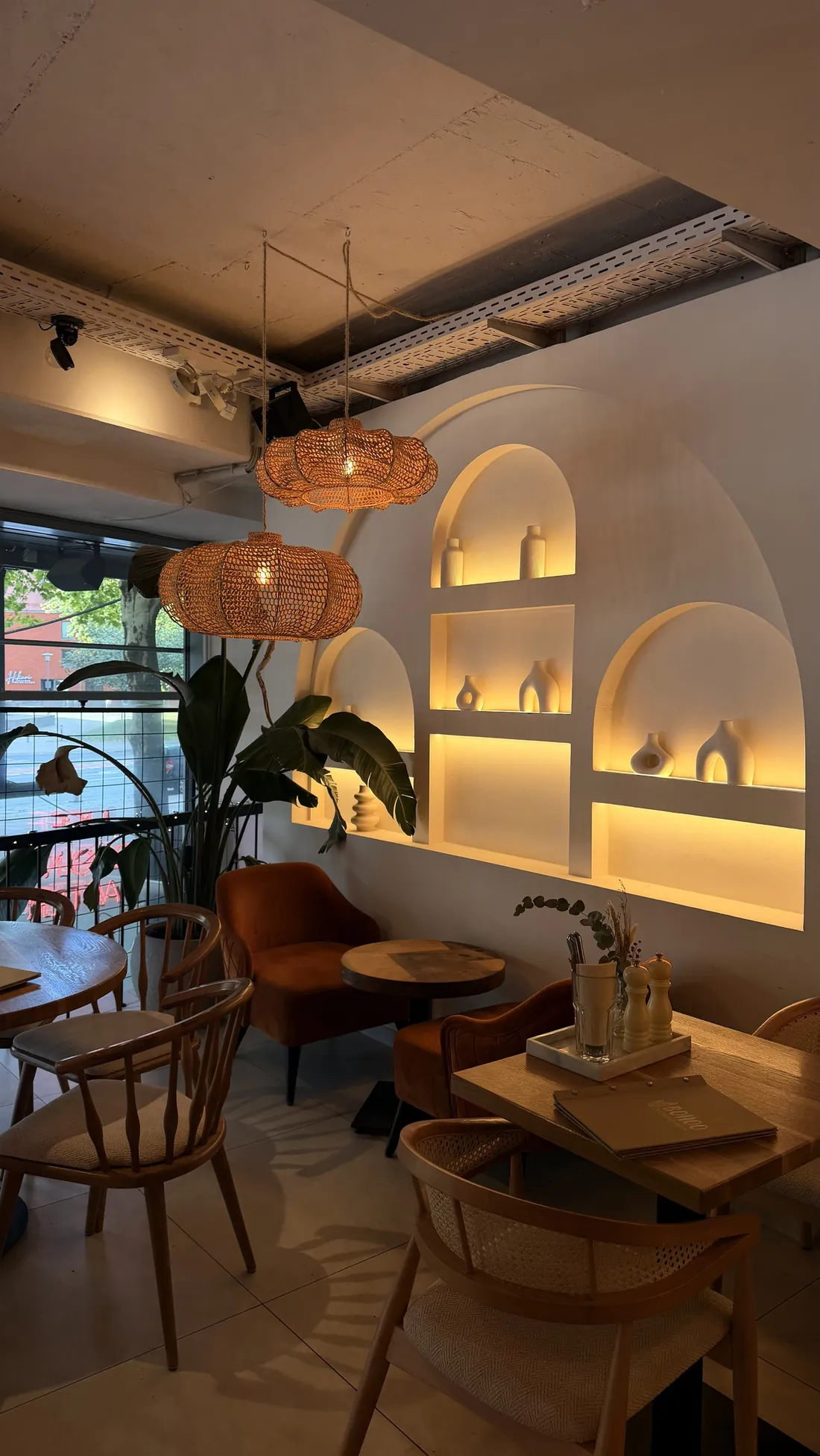 Lit arched wall niches with white ceramic vases, two rattan lamps in front, a wooden table with Viennese cane chairs and a velvet armchair