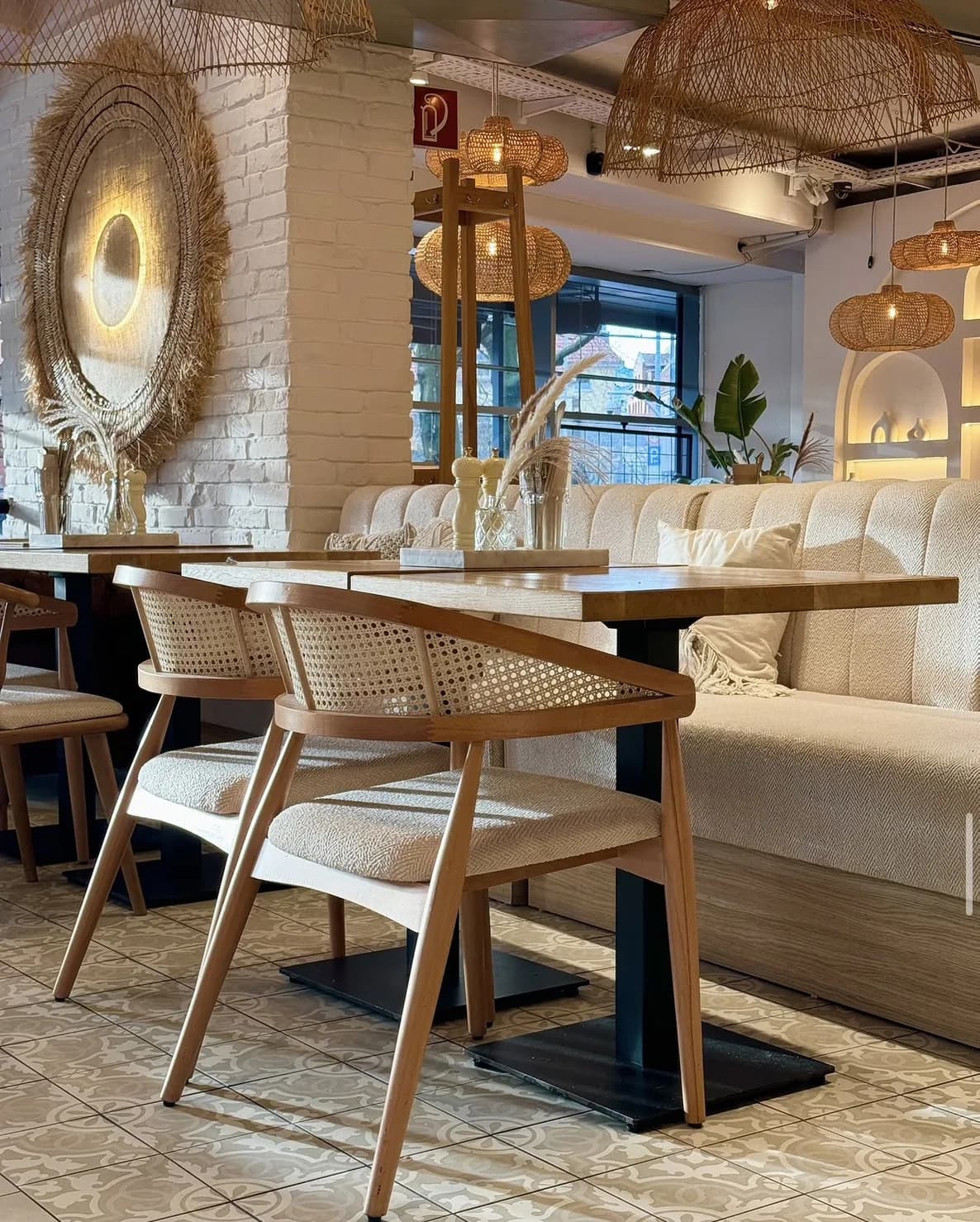 Inside Café Bohoo: a light upholstered bench, wooden chairs with Viennese cane and rattan lamps in front of a white brick wall
